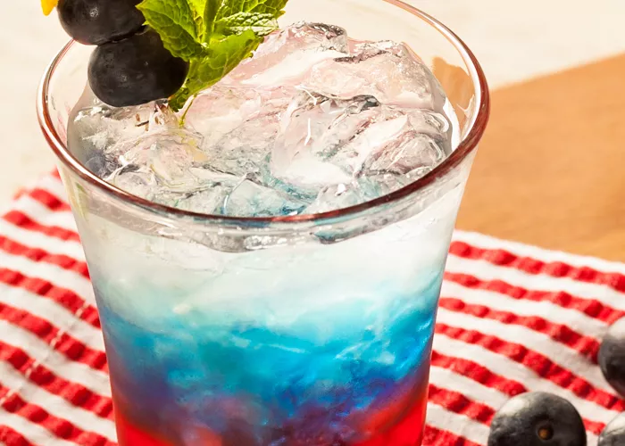 Glass of Citrus-Spangled Spritzer garnished with mint leaves, lemon rind star and berries alongside some blueberries  on red white striped cloth