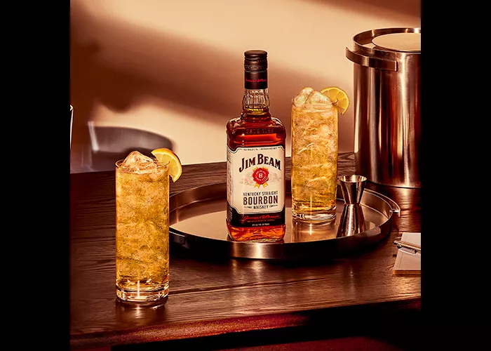 Bottle of Jim Beam  with a glass of Jim Beam® and Ginger Highball  along with measuring flask  in a plate  and a glass of Jim Beam® and Ginger Highball with a ice bucket on the table 