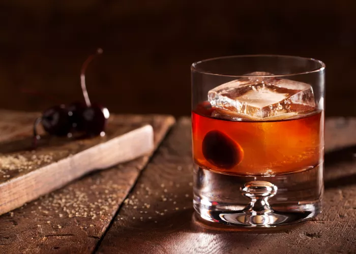 A glass of Classic Old Fashioned, garnished with a cherry and an ice cube, sits alongside cherries on a wooden plank placed on a wooden table.