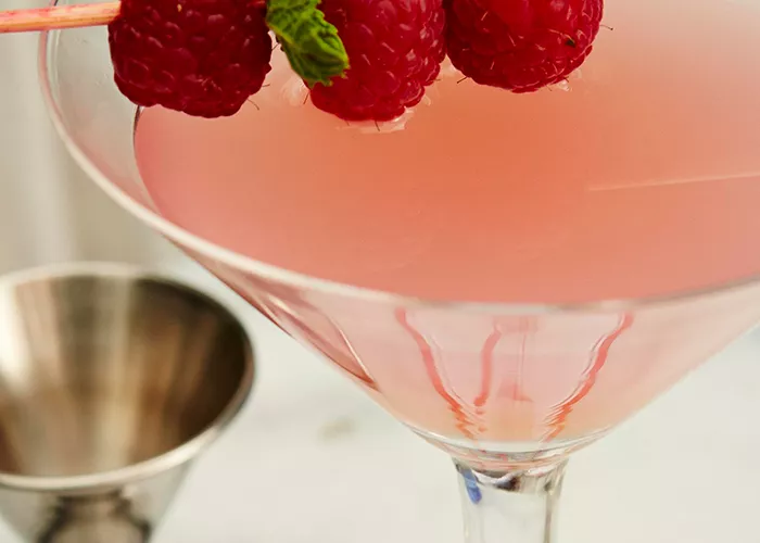 A cocktail glass of Paris Escape garnished with raspberries and lemon on a cocktail stick, accompanied by a jigger.