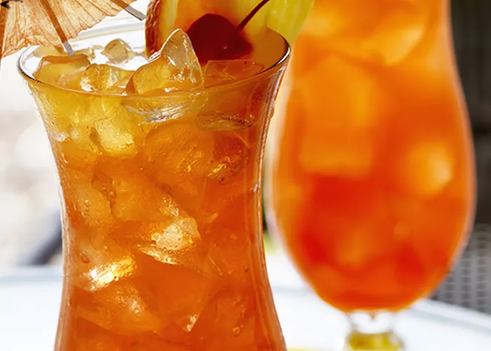 Two tulip glasses of Island Breeze garnished with pineapple slice, cherry, orange slice, with cocktail umbrella and couple of ice cubes.