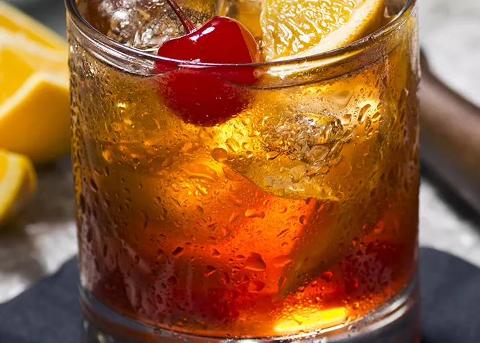 A mug of New Fashioned garnished with cherry and lemon slice  with some orange slices placed besides.