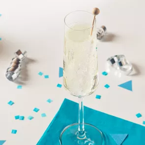 A flute glass of Midnight's Kiss, garnished with a rock candy stick, rests on a blue paper cloth, accompanied by small pieces of paper and a silver spiral sheet on the table.