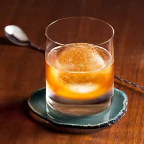 A glass of Maple Leaf placed on a coaster garnished with Scrape fresh cinnamon on top . Along with steel spoon placed on wooden slab.