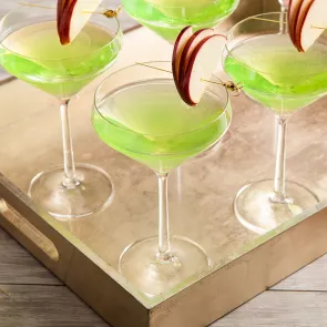 Four glasses of Appletini, each garnished with an  apple slice pierced on a cocktail pick on the rim  of the glass, are arranged in a wooden tray.