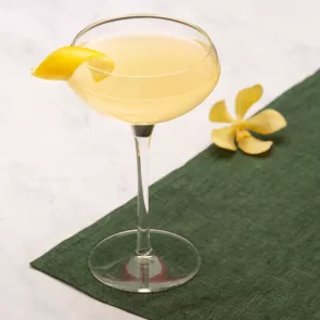 A glass of Bee's Knees garnished with lemon peelon a green cloth alog with the yellow flowers  and bowl of lemons 