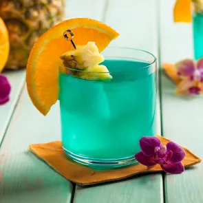  Glass of Blue Hawaii garnished with an orange wedge and a pineapple in pick on a orange napkin alongside a pink orchid