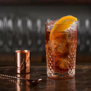 A glass of Negroni, garnished with an orange slice and ice cubes, is accompanied by a copper spoon and a copper glass.