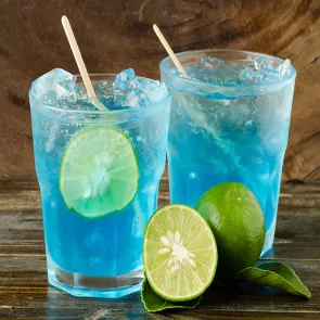 Two glasses of Home Team Punch with ice cubes, lime slice and stirrer in it alongside one and half  lime with leaves