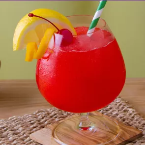 A snifter glass of Sunset Daiquiri placed on a coaster  and jute mat, garnished with cherry, lemon slice, lemon peel with straws inside.