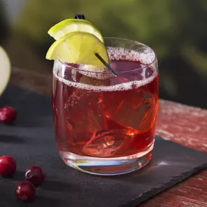 A glass of Orchard Twist garnished with lemon slice containing crushed ice and accompanied by a few cranberries on a black tray.