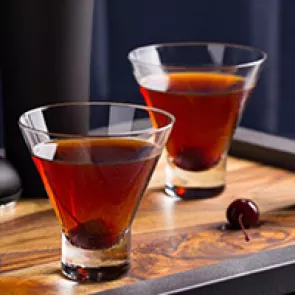 Two glasses of Jim Beam Black Manhattan garnished with a cherry in each, alongside a cherry on a wooden tray.