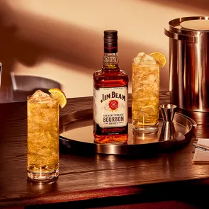 Bottle of Jim Beam  with a glass of Jim Beam® and Ginger Highball  along with measuring flask  in a plate  and a glass of Jim Beam® and Ginger Highball with a ice bucket on the table 