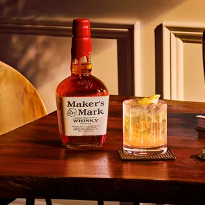 Glass of Maker's Mark® Gold Rush on a coaster alongside bottle of Maker's Mark Whiskey