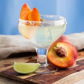 A glass of Sauza® Peach Margarita, garnished with wedges of peach, accompanied by a slice of lime and a slice of peach, is placed on a wooden tray