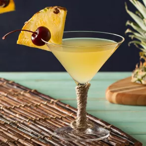 A glass of Pina Colada Martini garnished with a slice of pineapple and a cherry on the rim, placed on a wooden mat