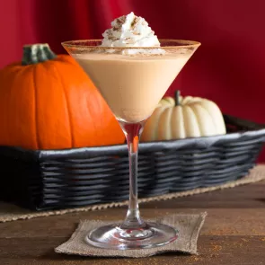 A glasses of Pumpkin Pie Martini garnished with whipped cream and Pumpkin Spice, placed on a small piece of cloth. Nearby, a tray of pumpkin on a cloth