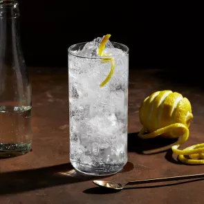 A glass of Roku® Gin Sonic with ice, half-filled water bottle, half-filled green glass, artisticallly chopped lemon, and copper spoon on the table.