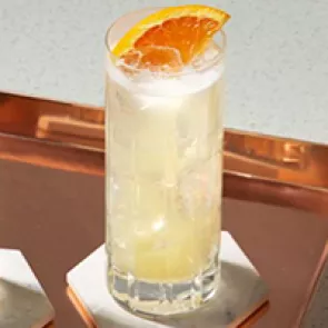 A  glass of Tom Collins garnished with chopped orange in a copper plate on a stone coaster 