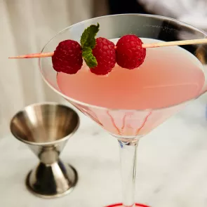 A cocktail glass of Paris Escape garnished with raspberries and lemon on a cocktail stick, accompanied by a jigger.