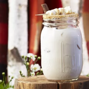 A jar of S'mores, garnished with marshmallows on a wooden rock, with a cocktail stick, chocolate stick and sugar rim.