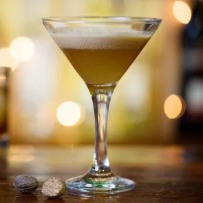 A cocktail glass of A Warm Welcome with fresh nutmeg, sprinkle on top for garnish with pieces of nutmeg alongside it.