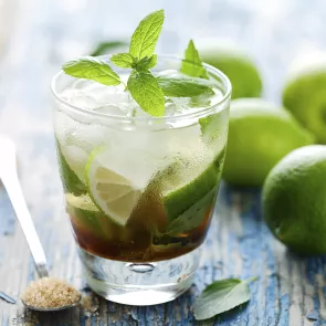 A glass of Banana Cinnamon Mojito garnished with fresh spanked mint and some lemon slices. alongside few lemons, some mint leaves and a steel spoon filled with sugar.