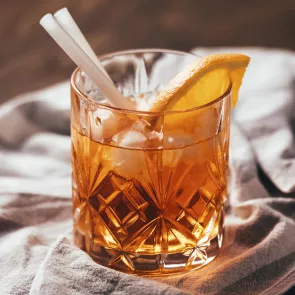 A glass of Coconut Old Fashioned with coconut water ice cubes, garnished with an orange slice on the rim, and two straws