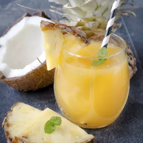 A glass of Delusion garnished with a piece of pineapple and few mint leaves along with a straw alongside a halved coconut and  a pineapple.