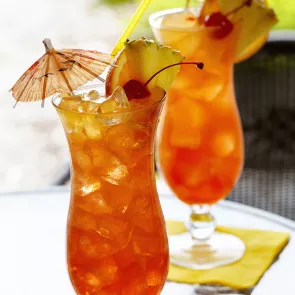 Two tulip glasses of Island Breeze garnished with pineapple slice, cherry, orange slice, with cocktail umbrella and couple of ice cubes.