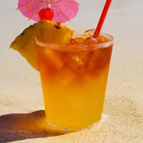 A glass of Mango Hurricane, garnished with an orange wedge and a cherry, along with a pineapple slice and a cocktail umbrella on the rim of the glass with a straw.