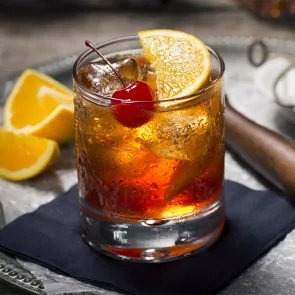 A mug of New Fashioned garnished with cherry and lemon slice  with some orange slices placed besides.