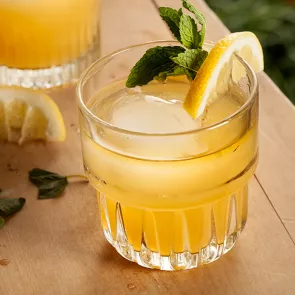 A glass of Nil Fashioned garnished with a slice of lemon and mint leaves, some of which are fallen on the wooden table.