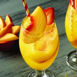 A glass of Peach on the Beach garnished with a peach slices and half cut strawsberry with staw. alongside a bowl of sliced peaches.