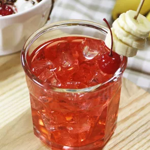 A glass of DeKuyper® Banana Split, garnished with ice cubes, a cherry, and banana slices pierced on a toothpick, is placed on the rim of the glass