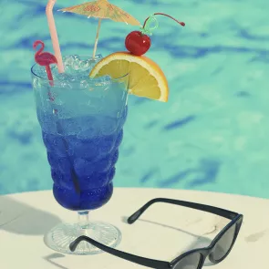 Glass of Blues on the Rocks with ice and a straws, garnished with a little umbrella, flamingo pick and a cherry and an orange slice on a pick