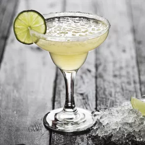 A glass of Elderflower Margarita garnished with ice cubes and a wheel of lemon on rim of glass