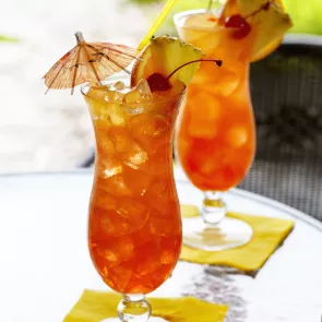 Two glass of Mai Time, garnished with ice cubes, a cherry, orange and  pineapple slice on the rim of the glass, and a cocktail umbrella