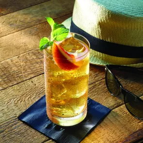 A glass of Peach Whiskey Sour with ice garnished with orange slice and mint leaves on black coaster alongside a sunglass and a hat