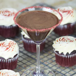 A glass of Red Velvet Cake Martini garnished with sugar mixture on rim of glass along side few cupcakes