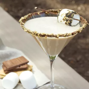 Glass of Smore-Tini garnished with a toasted marshmallow and rimmed with crushed graham crackers and melted chocolate, alongside some chocolate and marshmallows.