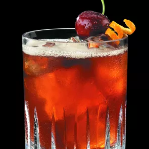 Glass of The Chee Chee with ice, garnished with an Amarena cherry and orange Zest.