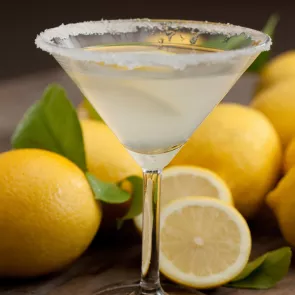 A martini glass of Kiwi Zest Martini, garnished with a lemon wedge and rimmed sugar. Alongside couple of lemons, lemon slices, and leaves.