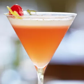 A martini glass of Pink Pineapple-Tini garnished with a cherry and pinapple sticks on a cocktail pick.