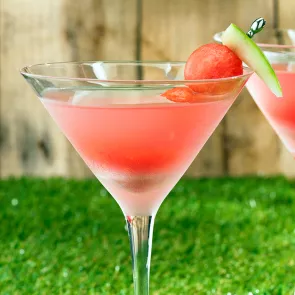 A martini glass of Pinnacle® Summer Breeze garnished with a watermelon wedge on cocktail pick placed on a green grass surface.