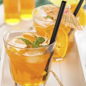 Two glasses of Sunset Tea garnished with an orange slice or apple spear dipped in cinnamon and mint leaves along with a straws and cocktail umbrella.