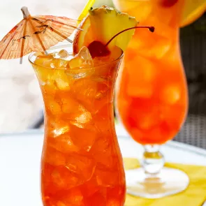 A glass of Watermelon Mai Tai, garnished with ice cubes, a cherry, orange and  pineapple slice on the rim of the glass, and a cocktail umbrella