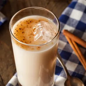 A glass of Horchata, spoon and few cinnamon sticks on blue & white checkered cloth, garnished with cinnamon and sugar.