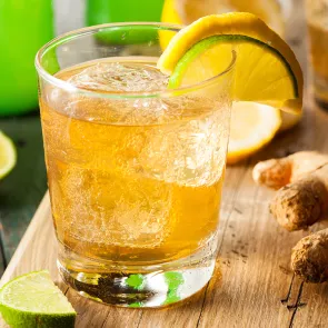 "A Glass of Mexican mule garnished with yellow/green lemon slices on glass rim and ice cubes inside along side ginger and slice of lemon placed on wooden table "