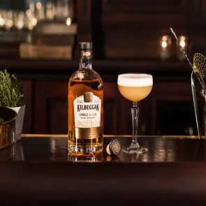 A bottle of Kilbeggan® Brosna Sour, accompanied by a glass of Kilbeggan® Brosna Sour, is placed on a black table.